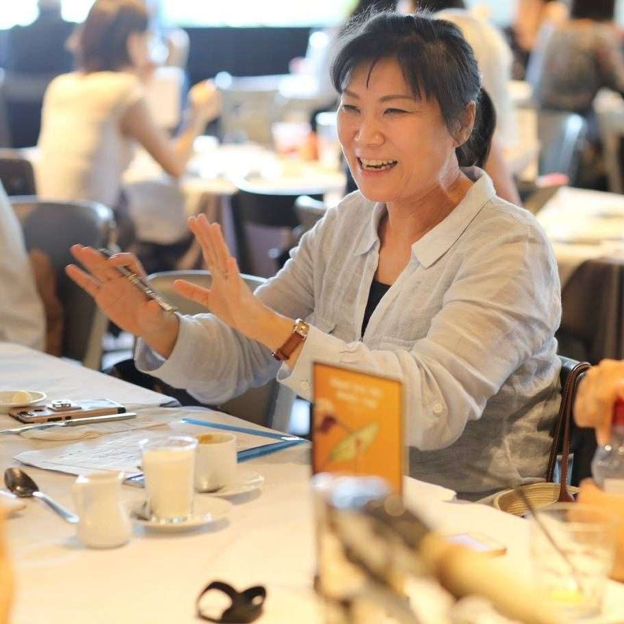 Smiling East Asian woman at a table, gesturing with her hands.