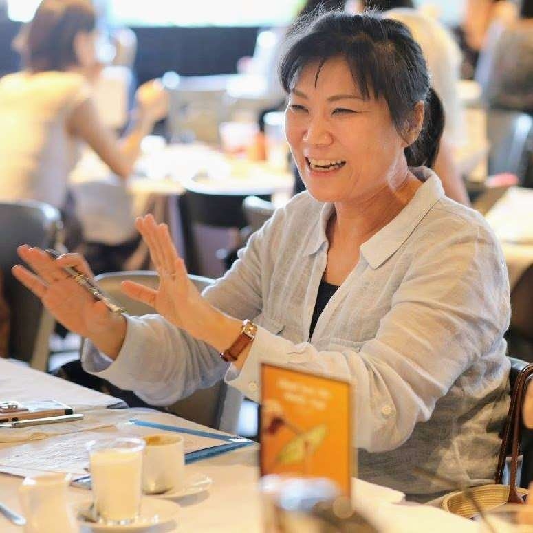 Smiling woman with black hair gesticulating with both hands at a team meeting (others are not visible). Ceramic coffee cups are on the table.