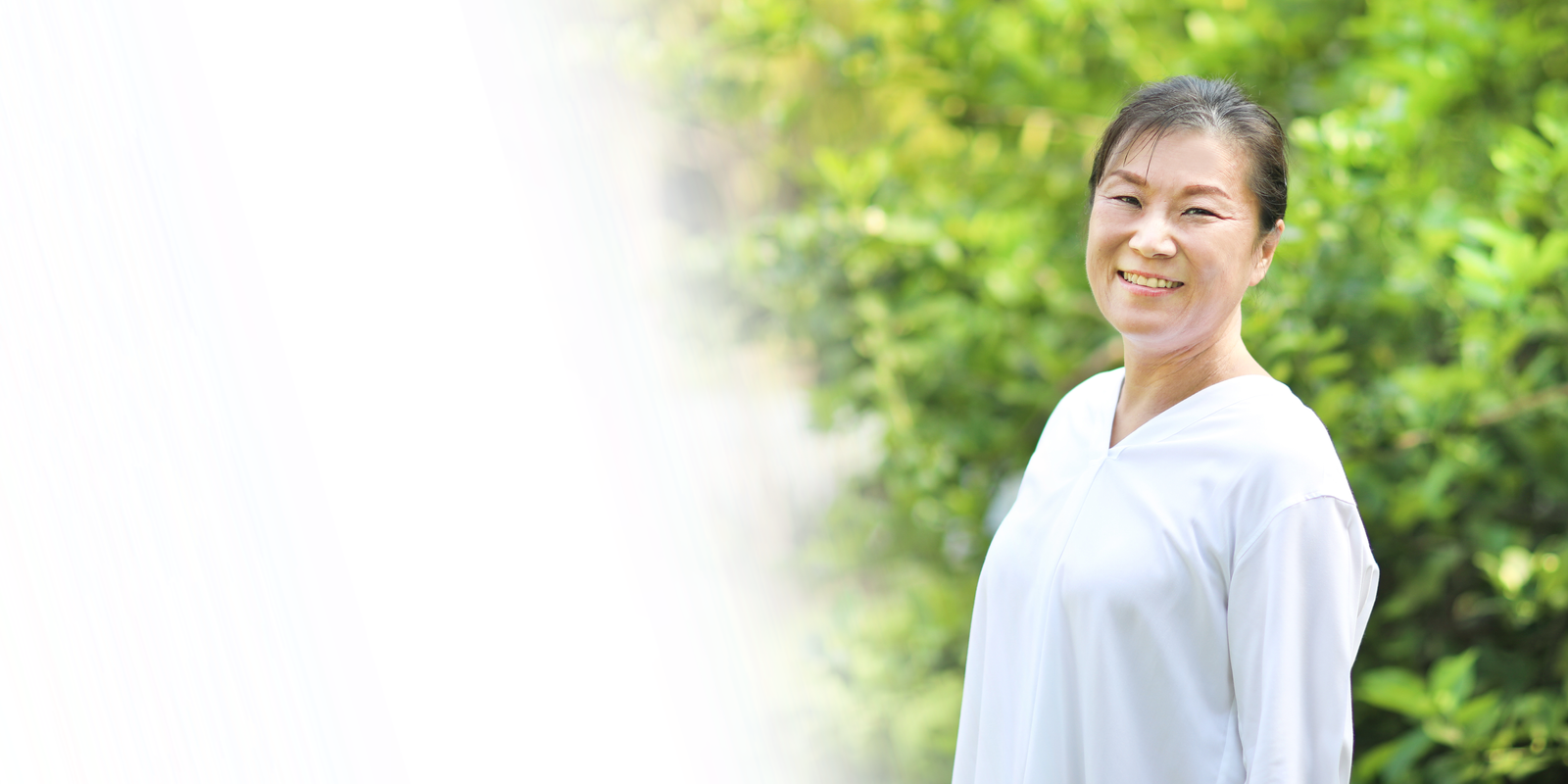Smiling East Asian woman in white shirt against green, leafy background.